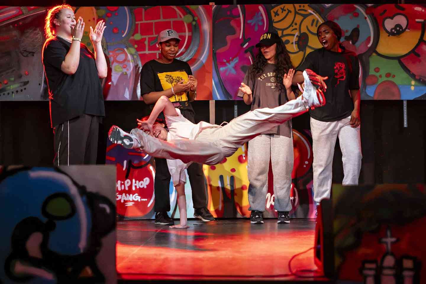 Eine Person führt auf einer Bühne vor buntem Graffiti-Hintergrund einen Breakdance-Move aus, während mehrere andere Personen daneben stehen und begeistert reagieren.