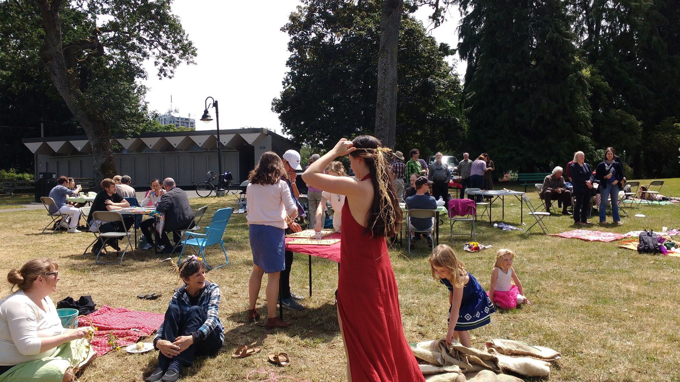 Menschen sitzen und stehen bei einem Picknick im Freien, einige auf Decken und andere an Tischen, während Kinder im Vordergrund spielen.