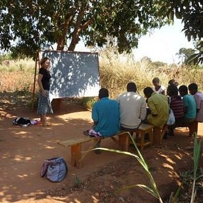 Gruppe von Menschen sitzt auf Bänken im Freien vor einer Tafel, eine Person unterrichtet unter einem Baum.