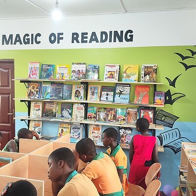 Kinder sitzen in einer bunt gestalteten Bibliothek in Ghana und lesen oder arbeiten, über den Bücherregalen steht der Schriftzug Magic of Reading.