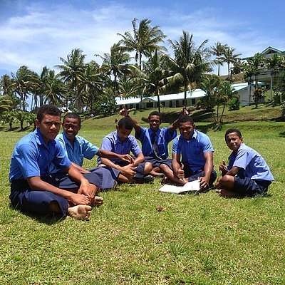 Eine Gruppe Jugendlicher in blauen Schuluniformen sitzt im Gras unter Palmen und arbeitet gemeinsam.