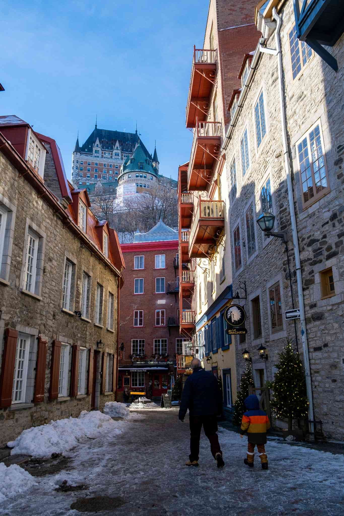 Zwei Personen gehen eine schmale Gasse mit alten Gebäuden entlang, im Hintergrund ein Schloss auf einem Hügel.