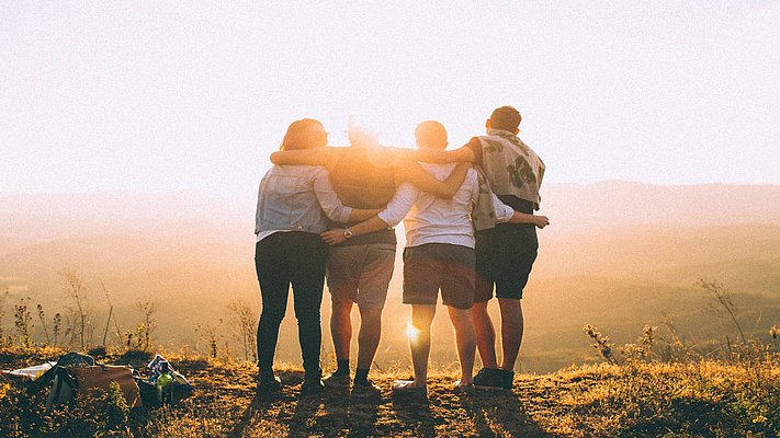 4 junge Menschen stehen mit dem Rücken zum Fotografen vor einer Landschaft