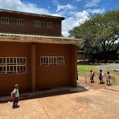 Kinder mit Rucksäcken laufen über den Hof einer Backsteinschule in Malawi.