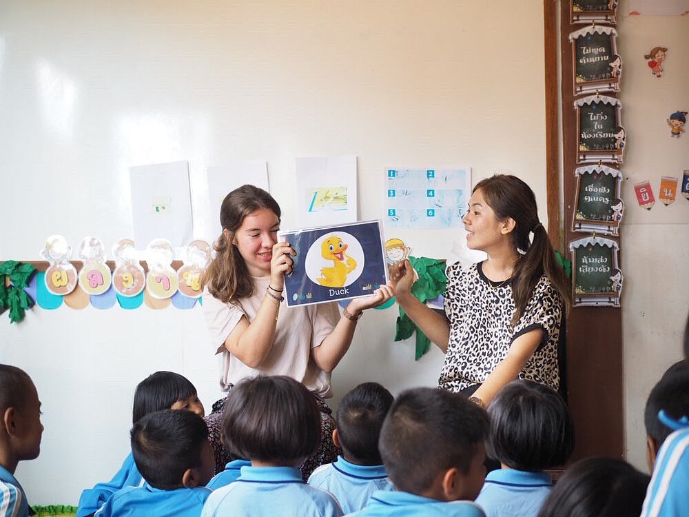 Zwei junge Frauen zeigen einer Gruppe von Kindern in blauen Schuluniformen ein Bild in einem Klassenzimmer.