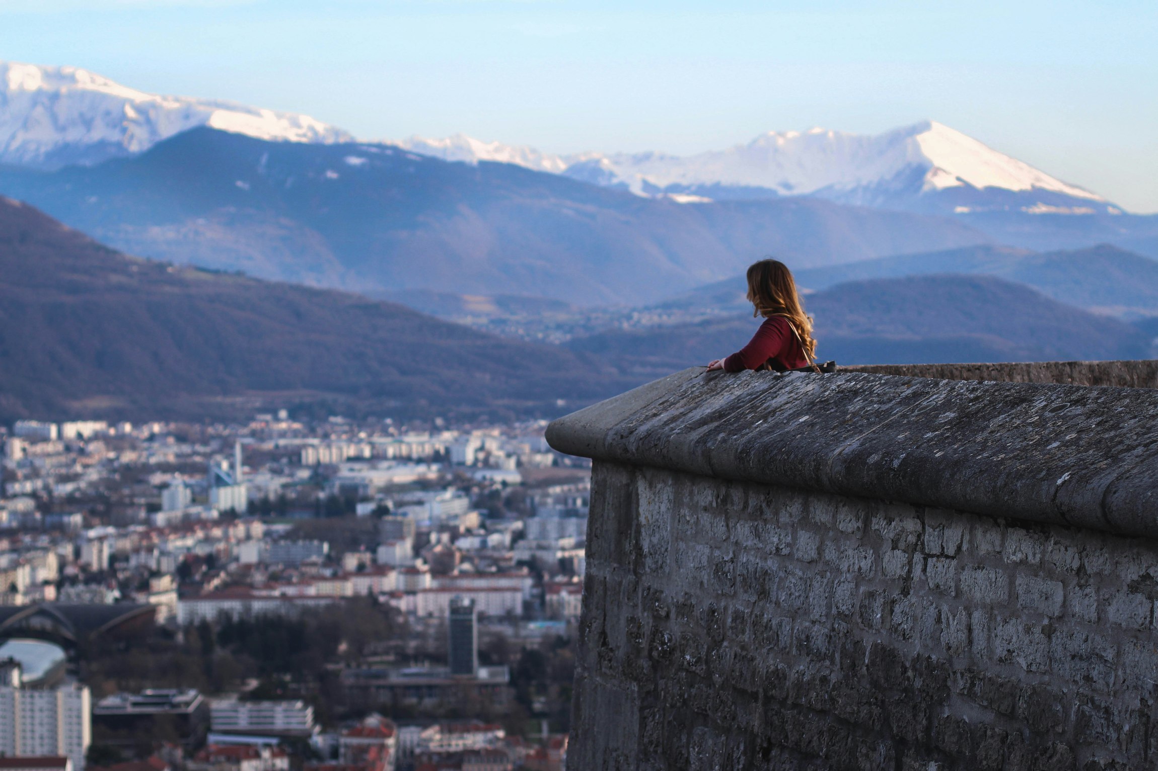 Person lehnt an einer Steinmauer und blickt auf Stadt mit Bergen und schneebedeckten Gipfeln im Hintergrund.