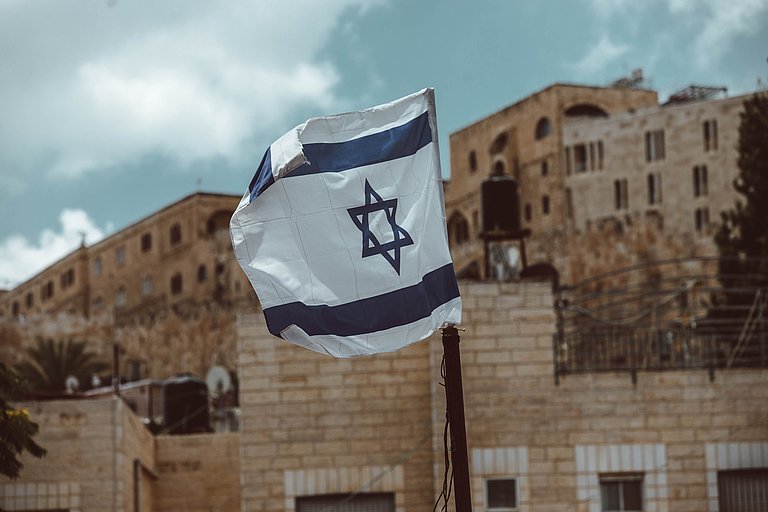 Israelische Flagge weht vor historischen Sandsteingebäuden unter bewölktem Himmel.