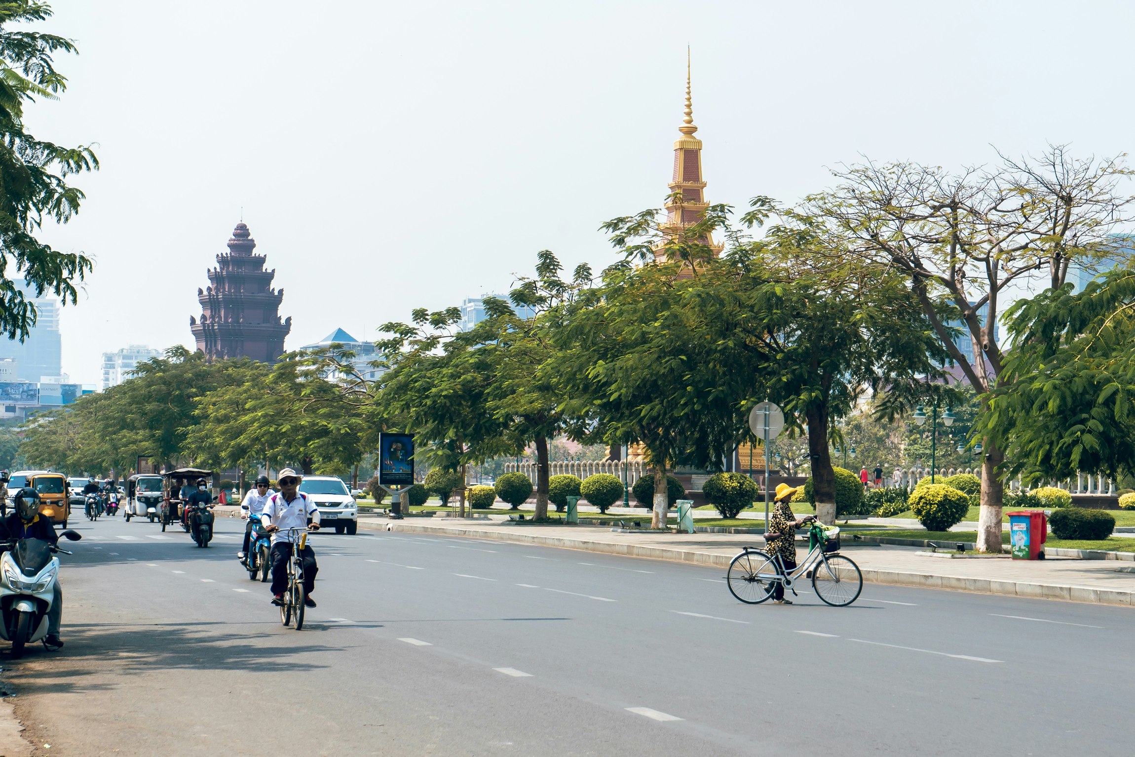 Breite Straße mit Autos, Motorrädern und Fahrrädern, im Hintergrund sind Pagoden und Bäume zu sehen.