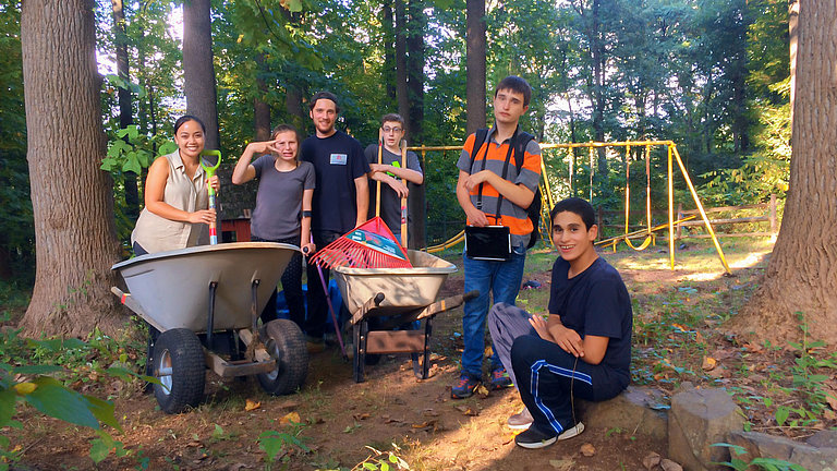 Eine Gruppe junger Menschen steht mit Schubkarren und Werkzeugen in einem Waldstück, im Hintergrund ist eine Schaukel zu sehen.