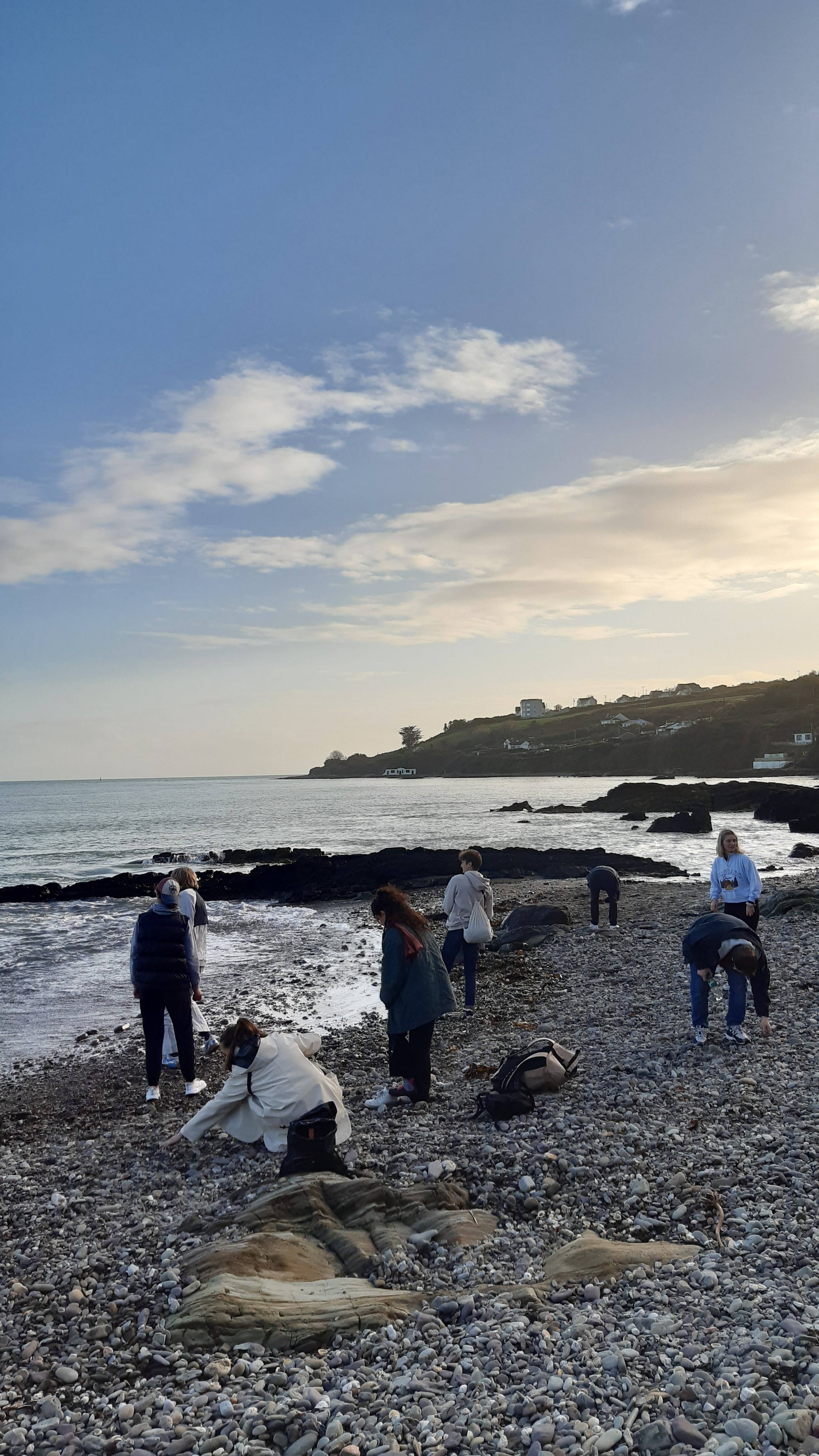 Mehrere Menschen sammeln Abfälle an einem steinigen Strand bei ruhigem Wetter.