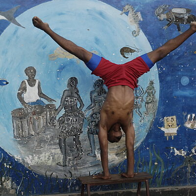 Person macht einen Handstand vor einer bunt bemalten Wand mit Trommlern und Fischen.