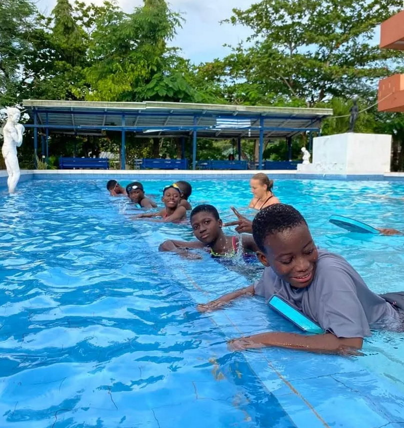 Kinder üben mit Schwimmbrettern am Beckenrand eines Pools, begleitet von einer Betreuungsperson.